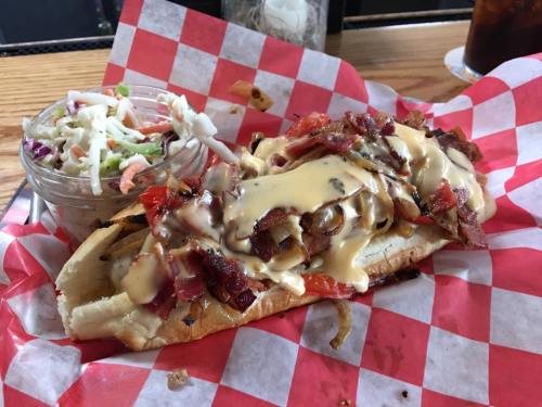 A loaded sandwich with bacon, cheese sauce, and toppings, served with a side of coleslaw in a glass bowl.