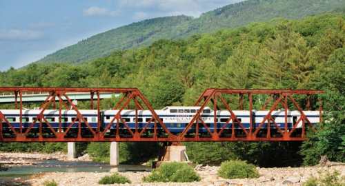 A train crosses a red metal bridge over a river, surrounded by lush green mountains and trees.