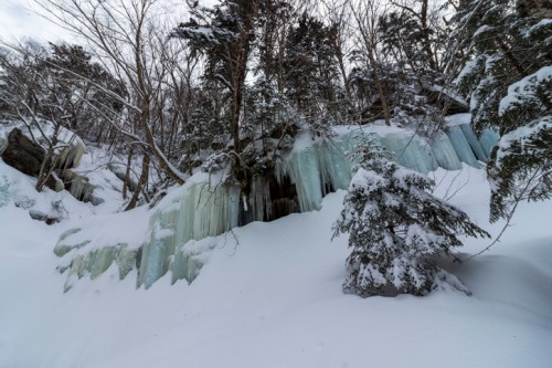 A snowy landscape featuring icy formations on a hillside, surrounded by bare trees and a blanket of fresh snow.