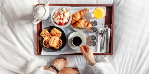 A breakfast tray on a bed featuring pastries, fruit salad, coffee, and orange juice, with a hand reaching for the cup.