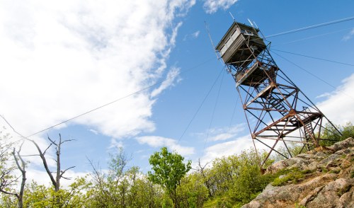 A tall fire lookout tower stands on rocky terrain under a blue sky with scattered clouds and greenery around.