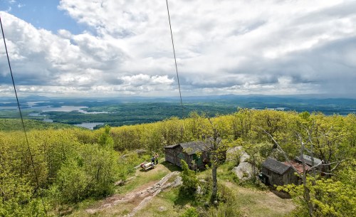 A scenic view from a mountain, featuring lush greenery, a small cabin, and a distant lake under a cloudy sky.