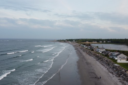 Aerial view of a sandy beach with gentle waves, lined by houses and greenery under a cloudy sky.