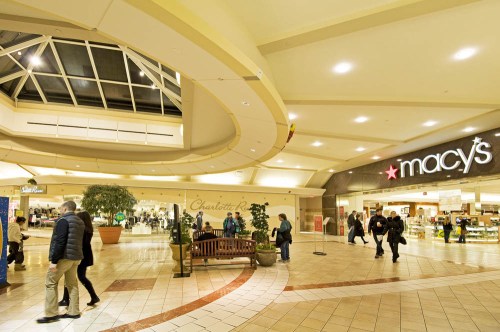 Interior of a shopping mall featuring a Macy's store, with shoppers walking and a seating area in the foreground.