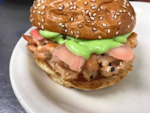 A sesame seed bun filled with a green sauce, sliced fish, and toppings, served on a white plate.