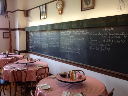 A cozy restaurant interior with checkered tablecloths, a chalkboard menu, and wooden chairs.
