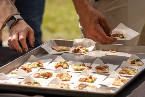 A tray of small appetizers with a hand placing one on a paper napkin, surrounded by more food items.