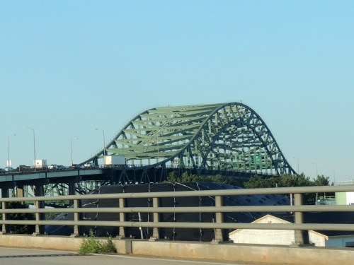 A green arch bridge spans over a highway, with vehicles traveling on it under a clear blue sky.