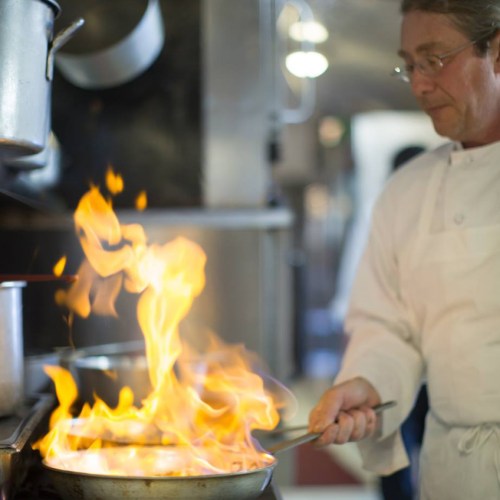 A chef in a white coat skillfully manages a flaming pan in a busy kitchen.