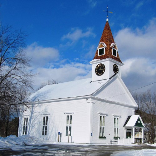 A white church with a clock tower and a snowy landscape under a blue sky with clouds.