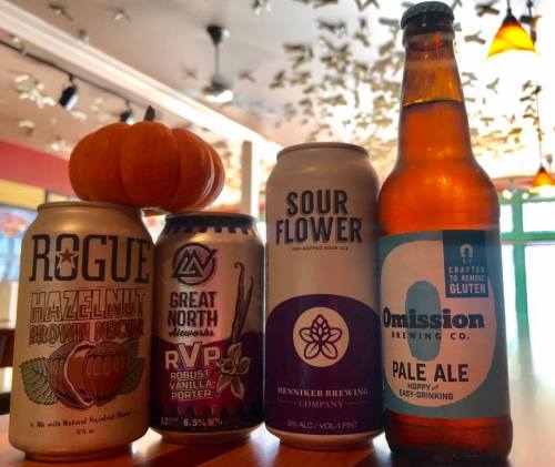 Four craft beer cans and a small pumpkin on a table, with a colorful background of hanging decorations.