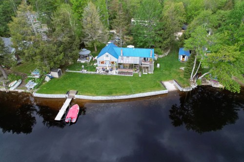 Aerial view of a lakeside property with a blue-roofed house, green lawn, and a pink boat docked by the water.