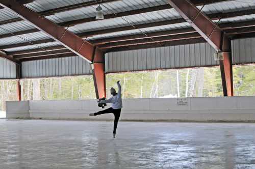 A figure skater performs a graceful move on an indoor rink with metal beams overhead.