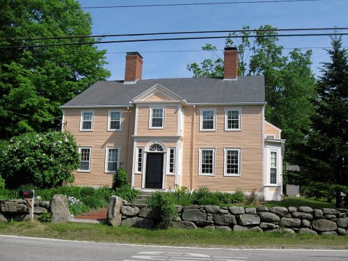A large, light peach-colored house with white trim, surrounded by greenery and a stone wall, on a sunny day.