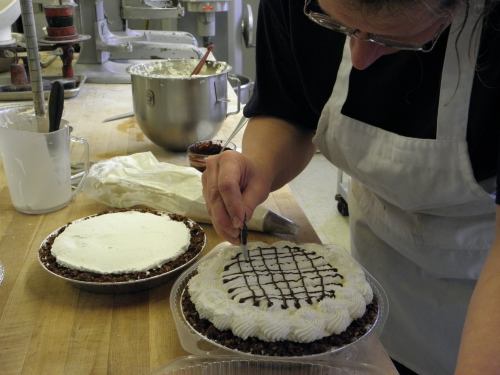 A baker decorates a pie with chocolate drizzle while another pie sits nearby on a wooden countertop.