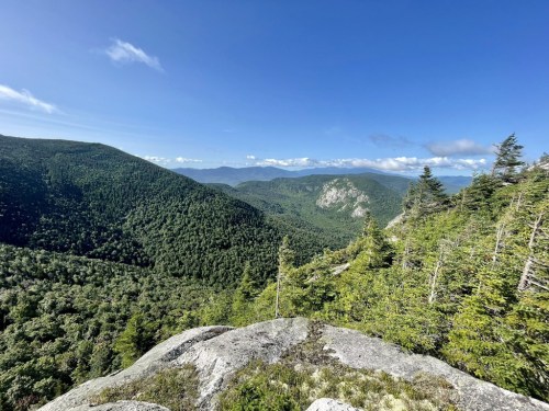 A panoramic view of lush green mountains under a clear blue sky, with rocky foreground and distant peaks.