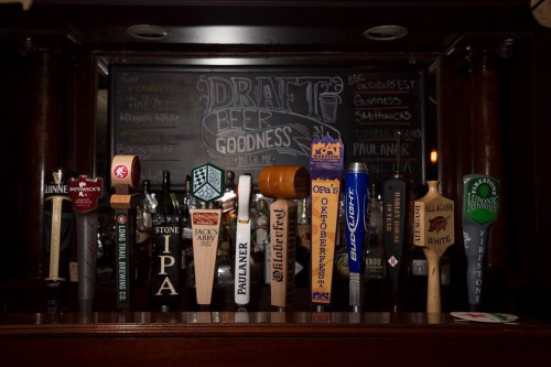 A variety of beer taps lined up on a bar, featuring different brands and styles against a chalkboard backdrop.