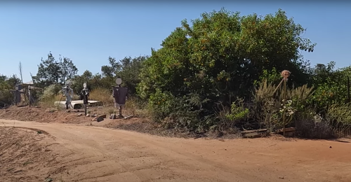 A dirt path leads through a rural area with bushes and two scarecrows standing among the greenery.