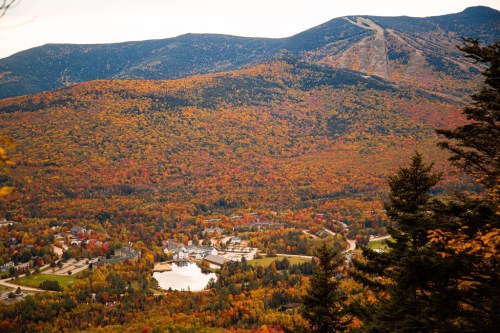 A scenic view of vibrant autumn foliage covering mountains and a small town, with a lake reflecting the colorful landscape.
