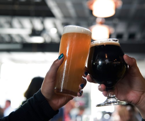 Two hands clink glasses of beer—one a light amber, the other a dark stout—against a blurred background of a lively bar.