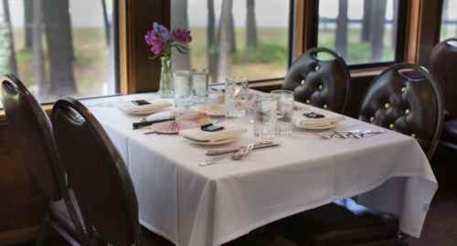 A neatly set dining table with a white tablecloth, glassware, and flowers, surrounded by wooden chairs by a window.