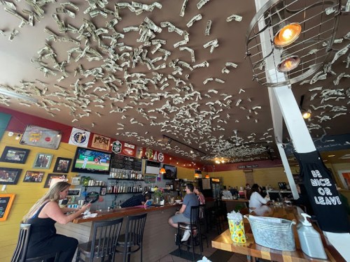 Interior of a lively restaurant with dollar bills covering the ceiling, bar area, and patrons enjoying their meals.