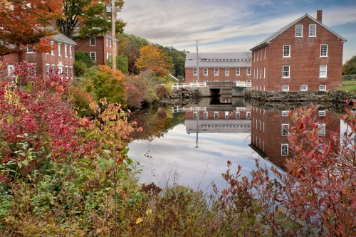 Historic red brick buildings reflect in calm water, surrounded by colorful autumn foliage and a serene landscape.