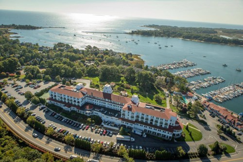 Aerial view of a waterfront hotel with a marina, surrounded by trees and a sparkling sea in the background.