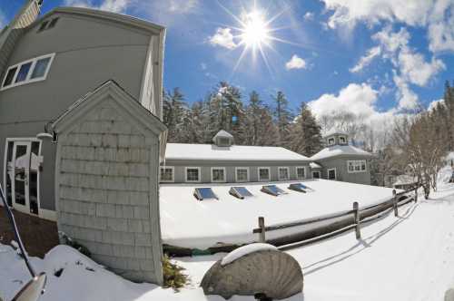 A snowy landscape featuring a gray building with solar panels, surrounded by trees and a bright sun in a blue sky.