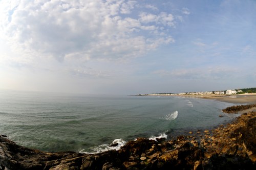 A serene beach scene with gentle waves, rocky shore, and a distant coastal town under a cloudy sky.