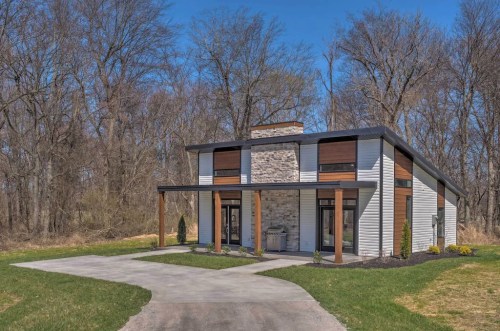 Modern two-story house with stone accents, large windows, and a driveway, surrounded by trees and greenery.