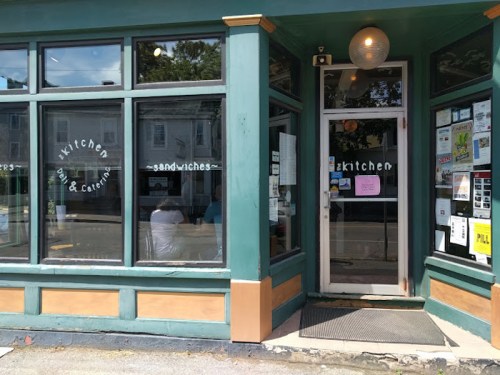 A quaint café entrance with large windows, featuring a sign that reads "kitchen" and "sandwiches."