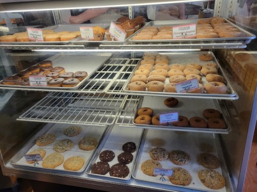 A display case filled with various donuts and cookies, labeled with names and prices.