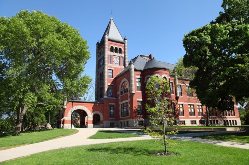 Historic red brick building with a clock tower, surrounded by green trees and a clear blue sky.