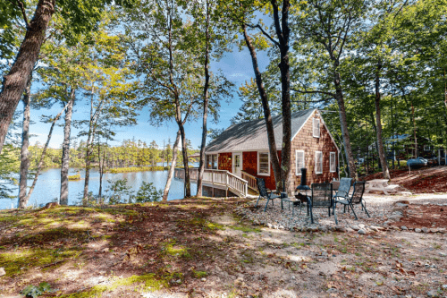 A cozy cabin by a serene lake, surrounded by trees and a small outdoor seating area.