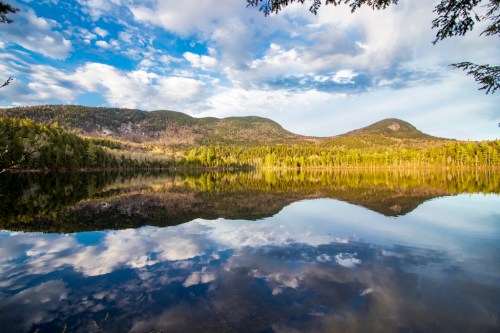 A serene lake reflecting mountains and a blue sky with clouds, surrounded by lush green trees.
