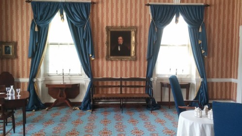 A room with patterned wallpaper, blue drapes, a portrait, and a wooden bench, featuring a table set with tea items.