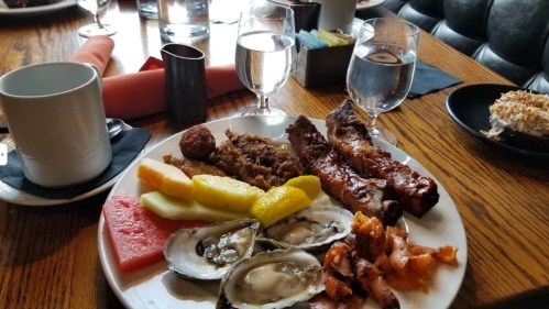 A plate of assorted food including ribs, oysters, fruit, and a dessert, with glasses of water on a wooden table.
