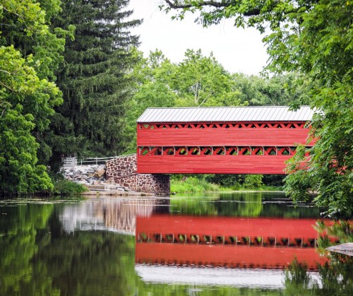 A red covered bridge spans a calm river, surrounded by lush green trees and reflecting in the water.