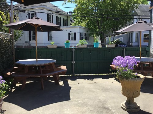 Outdoor seating area with picnic tables, umbrellas, and potted plants against a green fence and houses in the background.