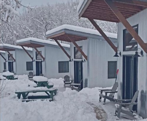 Snow-covered cabins with wooden porches and picnic tables, surrounded by a winter landscape of trees.