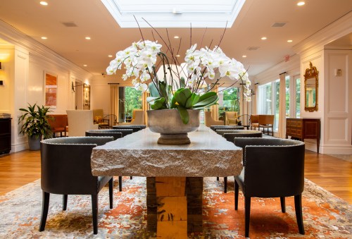 A modern lobby with a stone table, black chairs, and a large vase of white orchids, featuring warm wood floors and natural light.