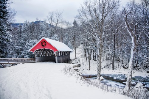 A red covered bridge in a snowy landscape, surrounded by trees and a stream, under a cloudy sky.