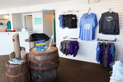 A retail space featuring clothing displays on walls, barrels, and a sign on a table, promoting shark-themed apparel.