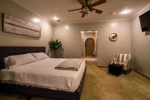 A cozy bedroom featuring a bed, armchair, and a doorway leading to a bathroom, with soft lighting and neutral colors.