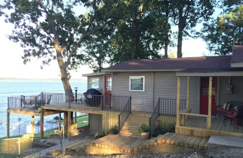A lakeside house with a wooden deck, surrounded by trees, overlooking the water on a sunny day.