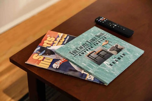 A coffee table with two magazines and a remote control on top. One magazine features sports, the other is a coffee table book.