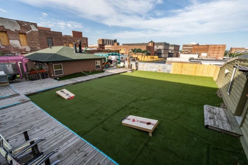 A spacious outdoor area with artificial turf, featuring two cornhole boards and surrounding buildings in the background.