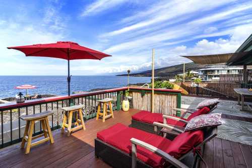 A scenic ocean view from a deck with red umbrellas, lounge chairs, and bar stools overlooking the water.