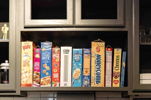 A variety of cereal boxes neatly arranged on a kitchen shelf, including Cheerios, Honeycomb, and Corn Flakes.